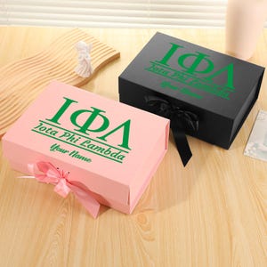 Può includere: Due scatole regalo, una rosa e una nera, sono esposte su una superficie di legno. Entrambe le scatole presentano le lettere greche IΦΛ e il testo "Jota Phi Lambda Your Name" in verde. La scatola rosa ha un nastro rosa, e la scatola nera ha un nastro nero.