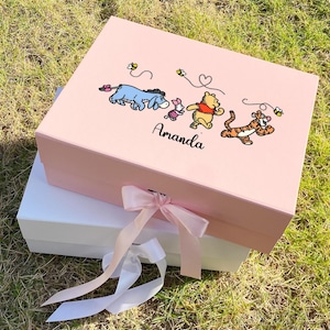 Peut inclure: Une boîte cadeau rose avec une base blanche, attachée avec un ruban rose. Le couvercle présente des illustrations de personnages de Winnie l'Ourson, des abeilles et le nom "Amanda" en écriture noire. La boîte est posée sur de l'herbe verte.