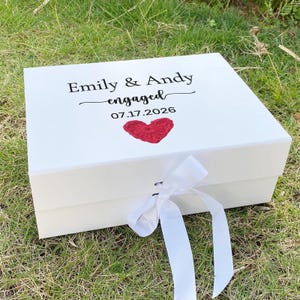 May include: A white rectangular gift box with a hinged lid and a white satin ribbon closure. The top of the box features the black printed text "Emily & Andy", "engaged", and the date "17.07.2026" above a small, textured red crocheted heart.