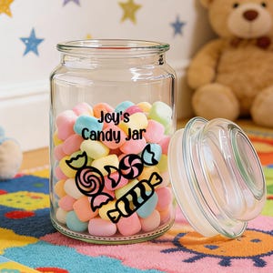 Peut inclure: Un bocal à bonbons en verre transparent rempli de bonbons aux couleurs pastel. Le bocal a un couvercle et porte l'inscription "Joy's Candy Jar" en noir. Des illustrations de bonbons noirs sont également présentes. Le bocal est posé sur un tapis coloré.