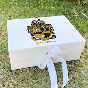 Może przedstawiać: Białe pudełko prezentowe z ozdobnym herbem i złotym napisem "Your Name". Herb przedstawia tarczę z czarnymi i złotymi elementami. Biała wstążka jest zawiązana wokół pudełka.