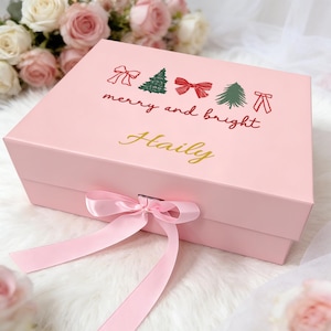 Puede incluir: Una caja de regalo rectangular de color rosa claro con tapa abatible y cierre de cinta rosa. La tapa presenta un gráfico de lazos rojos y árboles de Navidad verdes con el texto "merry and bright" y el nombre "Haily" en letra cursiva dorada.