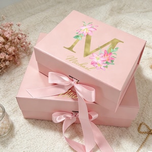 Puede incluir: Dos cajas de regalo rosas apiladas con lazos de cinta de satén. La caja superior presenta la letra dorada "M" con detalles florales y el nombre "Michael". Las cajas están sobre una superficie blanca.