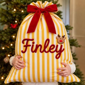 May include: A fabric gift sack featuring vertical yellow and white stripes, cinched at the top with a large red velvet bow. The name Finley is embroidered in red script, accompanied by a small, plush reindeer applique with a red nose.