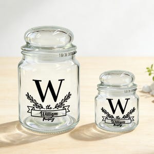 May include: Two clear glass jars with lids, one large and one small. Each jar features a black letter "W" with decorative leaves and the words "the William family" on a banner. The jars are on a light-colored wooden surface.