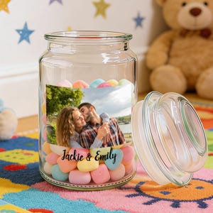 May include: A clear glass jar filled with colorful heart-shaped candies. A photo of a couple is adhered to the jar, with the names "Jackie & Emily" printed below. The jar's lid is off to the side, and the jar sits on a colorful rug.