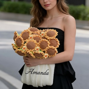 May include: A bouquet of crocheted sunflowers with brown centers, yellow petals, and green leaves. The bouquet is held in a cream-colored knitted bag with the name "Amanda" in script.
