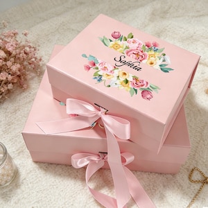 Peut inclure: Deux boîtes cadeaux roses empilées, ornées de compositions florales et du nom "Sophia" imprimé sur la boîte supérieure. Chaque boîte est ornée d'un ruban de satin rose noué en nœud. Les fleurs sont roses, jaunes et blanches.