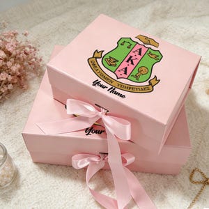 Op de afbeelding: Twee roze geschenkdozen met een strik. De bovenste doos heeft een wapen met Griekse letters en de tekst "Your Name". Het wapen is groen, roze en goud. De dozen zijn op elkaar gestapeld.