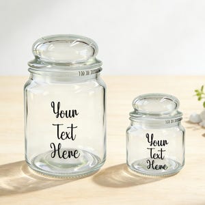 Op de afbeelding: Twee doorzichtige glazen potten met afgeronde deksels. De grotere pot heeft een bredere basis dan de kleinere. Beide potten hebben de tekst "Your Text Here" in zwart script. De potten staan op een lichtgekleurd houten oppervlak.