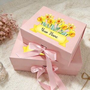 Puede incluir: Dos cajas de regalo rosas apiladas una encima de la otra. La caja superior tiene un diseño floral de narcisos amarillos y una pancarta amarilla con el texto "Your Name". Cada caja tiene una cinta rosa atada con un lazo.