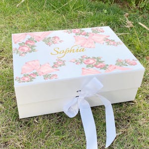 May include: A rectangular white gift box featuring a floral pattern of pink roses and light pink bows printed on the lid. The name Sophia is written in gold cursive script on the centre of the lid. A white satin ribbon is tied in a bow at the front.