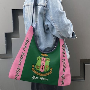 May include: A green and pink tote bag with the text "Alpha Kappa Alpha" in pink on the sides. The center features a crest with the letters "AKA" and the words "Your Name". The bag is carried by a person wearing a denim jacket.