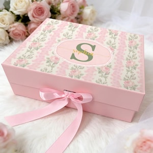 May include: A rectangular pink gift box with a hinged lid featuring a floral pattern of pink roses and green leaves. The center displays a scalloped oval label with a large green letter S and the name Sophia in gold script, tied with a pink ribbon.