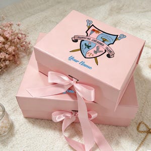 Peut inclure: Deux boîtes cadeaux roses empilées, ornées de rubans de satin assortis noués en nœuds. La boîte supérieure présente un écusson avec le texte "Founded 1945 Lewis Business College" et "Your Name".