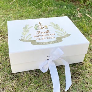 May include: A white rectangular gift box with a white ribbon closure. The lid features a watercolor illustration of a church, green olive branches, and the text: Jade BAPTIZED ON 02.25.2026 BATIZADO DO.
