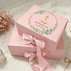 Peut inclure: Deux boîtes cadeaux roses avec des nœuds en ruban de satin. La boîte supérieure présente une croix dorée et un motif de couronne avec le texte "Jade Baptized on 02.25.2025". Les boîtes sont empilées l'une sur l'autre.