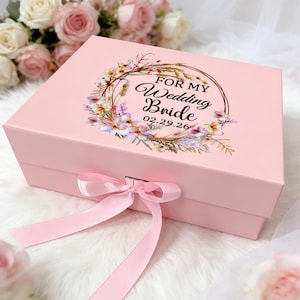 May include: A light pink gift box with a matching ribbon bow closure. The box lid features a floral wreath design with the text "FOR MY Wedding Bride 02.29.26". The box is on a white, fluffy surface, with a blurred floral background.