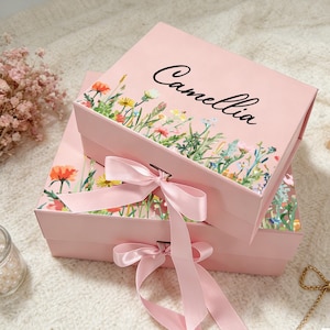 Può includere: Due scatole regalo rosa con disegni floreali e il nome "Camellia" stampato sulla parte superiore. Ogni scatola è adornata con un nastro di raso rosa legato a fiocco. Le scatole sono decorate con fiori selvatici colorati.