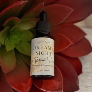 May include: A black dropper bottle with a white label featuring the text "DREAMY NIGHT Retinol Serum" and "1 oz by Volume." The bottle is set against a backdrop of red and green succulent leaves.