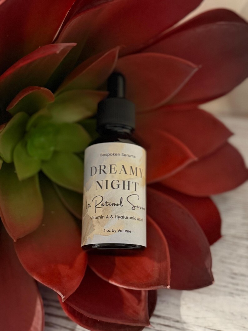 May include: A black dropper bottle with a white label featuring the text "DREAMY NIGHT Retinol Serum" and "1 oz by Volume." The bottle is set against a backdrop of red and green succulent leaves.