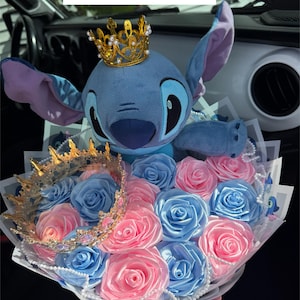 May include: A bouquet featuring a blue plush character wearing a gold crown, surrounded by pink and blue fabric roses. A second gold crown is also included. The bouquet is wrapped in white paper and accented with pearls.