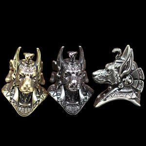 Puede incluir: Tres anillos de metal con la cabeza de Anubis, el dios egipcio. Los anillos están en acabados dorado, plateado y plomizo. Cada anillo tiene detalles intrincados, incluyendo la cara de perro y la tocado.