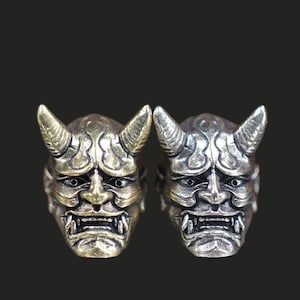 May include: Two rings with a detailed Oni mask design. One ring has a bronze finish, the other a silver finish. Each mask features horns, a stern expression, and sharp teeth. The rings are set against a dark background.
