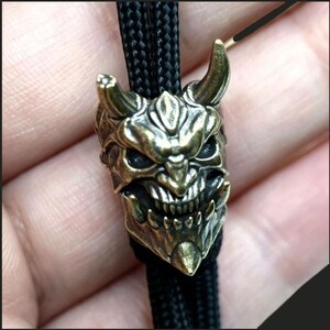 May include: A metallic brass-toned bead shaped like a horned demon mask with intricate facial features, including sharp teeth and deep-set eyes. The bead is threaded onto a black braided cord, serving as a decorative accent or functional pull.