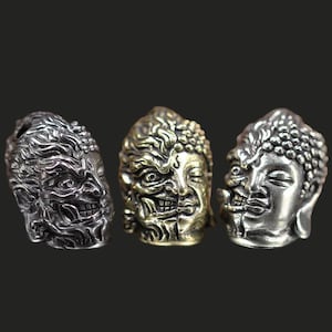 Puede incluir: Tres esculturas de cabezas de Buda de metal con un diseño único. Cada cabeza está dividida, con un lado que representa un Buda sereno y el otro una cara feroz y demoníaca. Las esculturas son en tonos plateados y dorados, sobre un fondo oscuro.