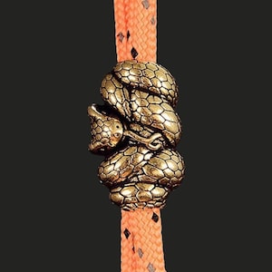 May include: A metallic gold-toned snake bead with a detailed scale texture coiled around a bright orange woven cord. The snake is depicted in a curled position with its head visible on the left side, featuring a smooth finish and dark accents.