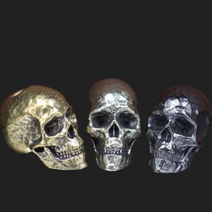 Könnte beinhalten: Drei Metallschädel-Figuren in Gold-, Silber- und Bronzetönen. Jeder Schädel hat detaillierte Gesichtszüge, einschließlich Augenhöhlen, einer Nasenhöhle und Zähnen. Die Schädel werden vor einem dunklen Hintergrund gezeigt.