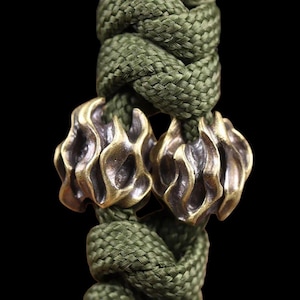 May include: A close-up shot of a green braided rope featuring two bronze-coloured, flame-shaped beads. The beads have a textured surface and are set against a black backdrop. The rope is a dark olive green colour.