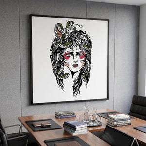 May include: A square wall art print featuring a black and white line drawing of a face with snakes for hair. The figure has red accents around the eyes and lips. The artwork is displayed in a thin black frame on a grey panelled wall above a desk.