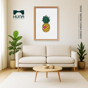 May include: A framed art print featuring a yellow pineapple with a pink heart, green leaves, and a white background. The print is displayed in a living room setting with a sofa, coffee table, and plants. Text on the right says "SIMPLY PRINT, FRAME, HANG."
