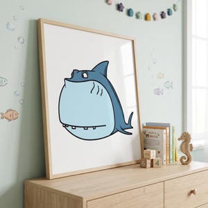 May include: A framed wall art print featuring a cartoon shark with a round, light blue body, a dark blue dorsal fin, and a wide mouth with visible teeth. The illustration is set against a plain white background within a light wood frame.