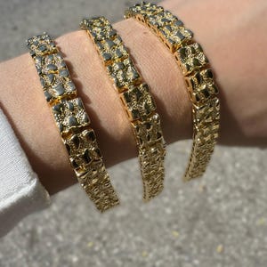 Può includere: Tre braccialetti dorati in stile pepita. Ogni braccialetto è composto da maglie rettangolari con una superficie strutturata. I braccialetti sono esposti su un polso, mostrando il loro design e colore.