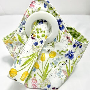 May include: A white fabric tote bag with a floral and bunny print. The bag features a large white circular handle and a green gingham lining. The print includes yellow tulips, blue irises, and white bunnies.