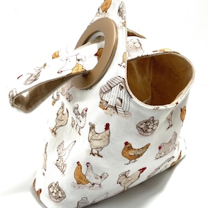 May include: A white fabric tote bag featuring a repeating pattern of chickens, chicken coops, and egg baskets. The bag has a brown handle and a circular opening. The interior is lined with a tan fabric.