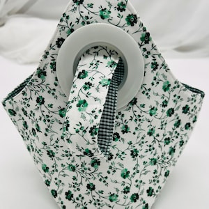 May include: A white fabric basket with a green floral pattern and a white handle. The interior and handle trim feature a black and white gingham pattern. The basket is designed to hold a roll of something.