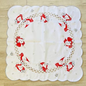 May include: A square white tablecloth with scalloped edges featuring a circular cutwork border. The fabric is decorated with embroidered red and white Santa Claus figures carrying gifts, interspersed with small grey embroidered stars.