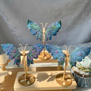Könnte beinhalten: Dekorative Schmetterlingsskulpturen mit blau-violetten Flügeln und goldfarbenen Ständern. Die Flügel weisen ein strukturiertes Muster auf. Die Schmetterlinge sind auf einer Holzoberfläche ausgestellt.
