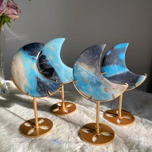 Pode incluir: Quatro esculturas de lua crescente com padrões marmorizados azuis, pretos e brancos. Cada lua é montada em um suporte de metal dourado com uma base circular. As esculturas são exibidas em uma toalha de mesa de renda branca.