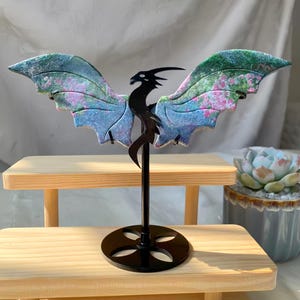 May include: A decorative dragon sculpture with outstretched wings. The wings are a blend of blue, green, and pink hues. The dragon's body is black and mounted on a black stand. The sculpture is displayed on a wooden surface.
