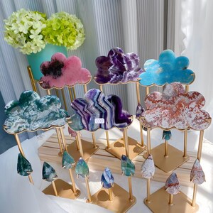 Pode incluir: Uma coleção de fatias de minerais em forma de nuvem montadas em suportes de metal dourado. Cada nuvem apresenta um padrão de pedra único em cores como rosa, roxo, azul, verde e vermelho, com pingentes de pedra em forma de gota pendurados.