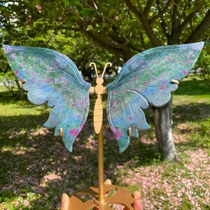 May include: A butterfly sculpture with blue and green wings, featuring pink accents. The wings are translucent. The butterfly is mounted on a gold-coloured stand. The sculpture is set against a blurred outdoor background.