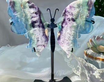 May include: A colorful butterfly sculpture with wings in shades of blue, purple, and green. The butterfly is mounted on a black metal stand. The wings are made of a translucent material, and the sculpture is displayed on a white surface.