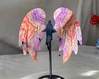 May include: A decorative angel wing sculpture. The wings are made of a polished stone with purple, orange, and white patterns. The wings are mounted on a black metal stand with a silhouette of a person in the center.
