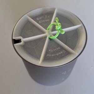 May include: A cylindrical grey container with a white plastic insert featuring a six-spoke divider. The insert has the text "Boil To Clean" and "Deadwood Gear Co" printed on it, with a bright green string loop attached to the centre point.