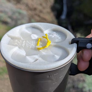 Puede incluir: Una taza de camping en blanco y negro con una tapa blanca. La tapa tiene un cordón amarillo adjunto. La taza tiene medidas en el lateral. El fondo es una vista borrosa de un paisaje natural.